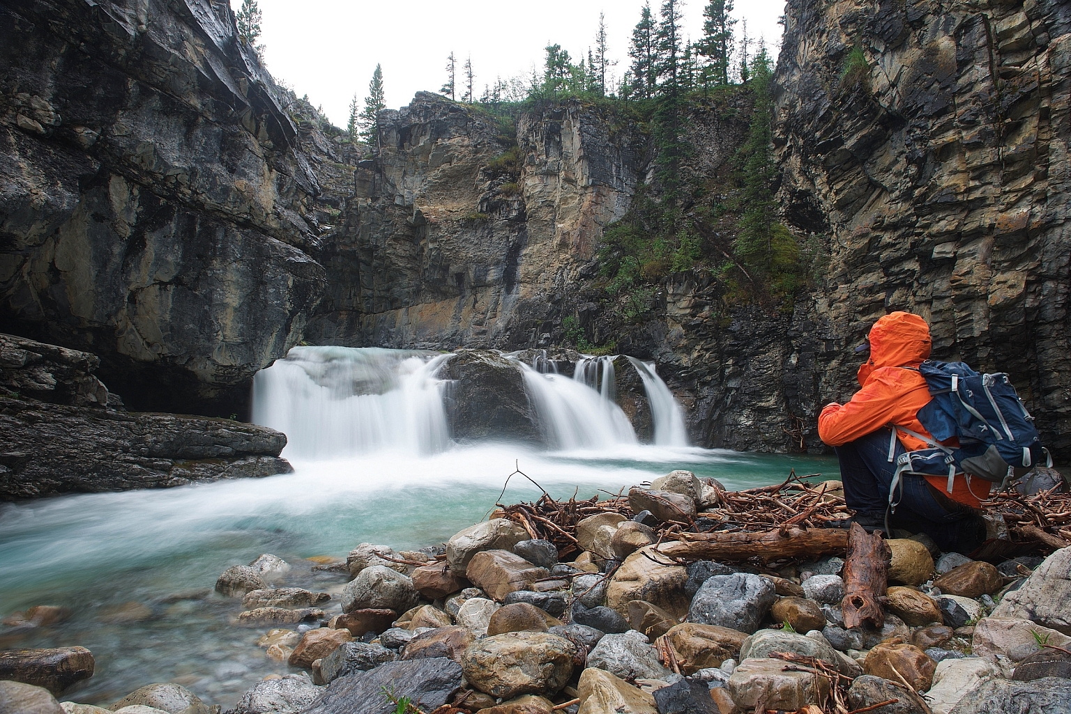 A person sits on a rocky shore looking out at a small waterfall in a canyon.