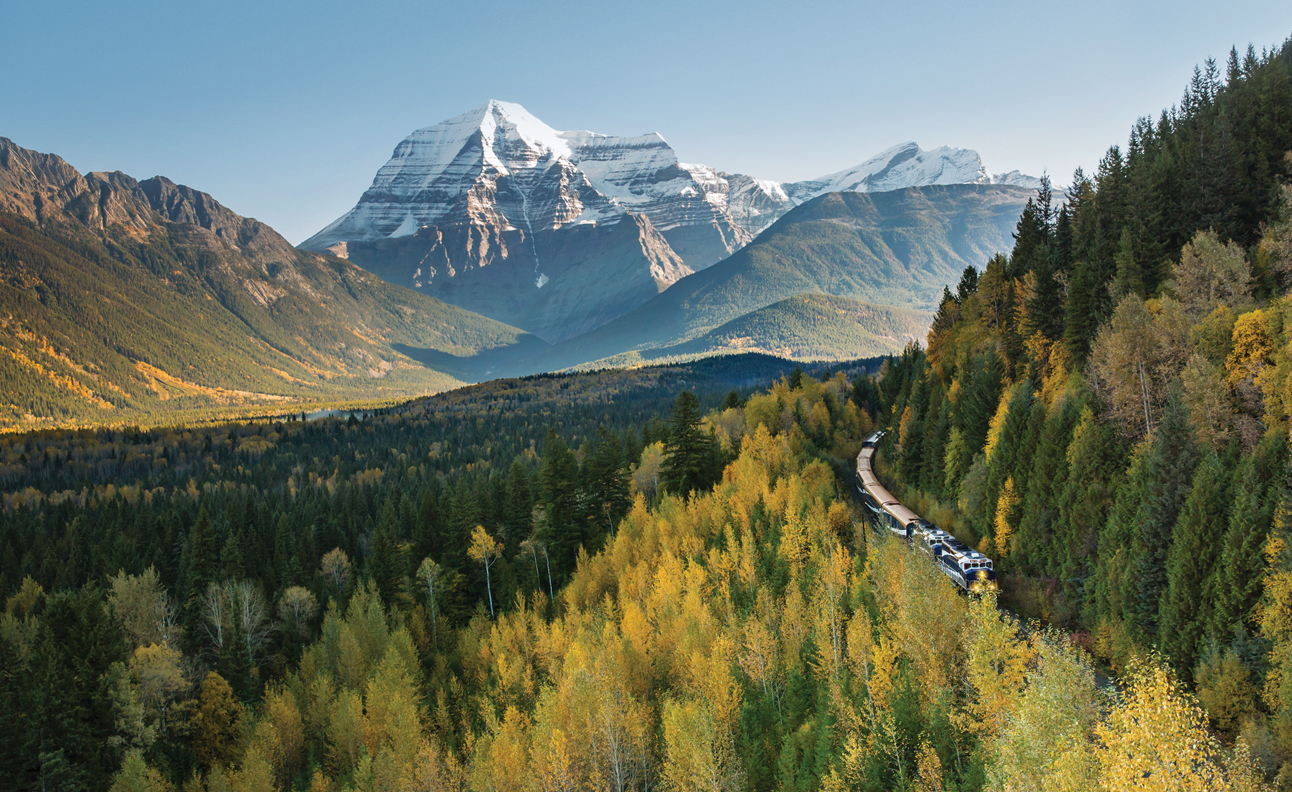 Aerial view of a train making its way along the side of a forested mountain, heading toward the camera. In the distance are snow-covered mountain peaks.